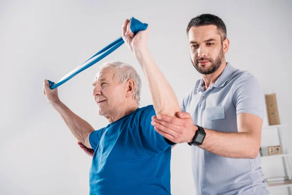 Vue latérale du thérapeute de réadaptation aidant l'homme âgé faisant de l'exercice avec du ruban en caoutchouc — Photo de stock