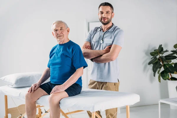 Terapeuta de rehabilitación con estetoscopio y hombre mayor en mesa de masaje - foto de stock
