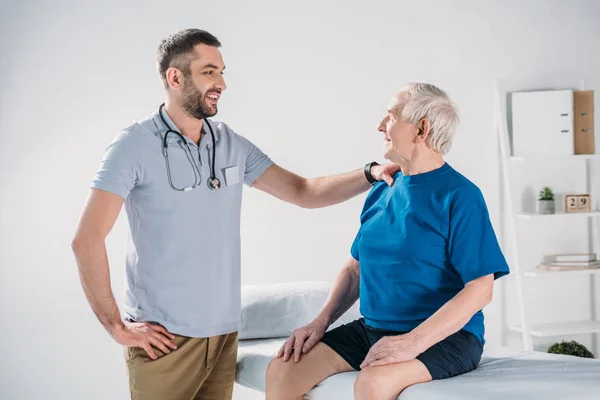 Rehabilitation therapist with stethoscope and smiling senior man on massage table — Stock Photo