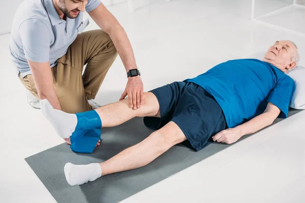Teilansicht eines Reha-Therapeuten, der einem älteren Mann beim Training mit Gummiband auf Matte hilft — Stockfoto
