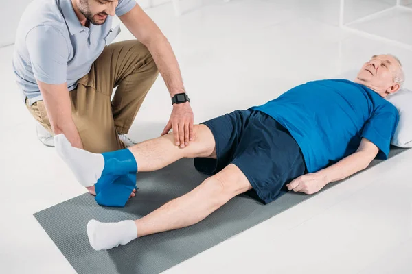 Vue partielle du thérapeute de réadaptation aidant l'homme âgé faisant de l'exercice avec du ruban de caoutchouc sur le tapis — Photo de stock