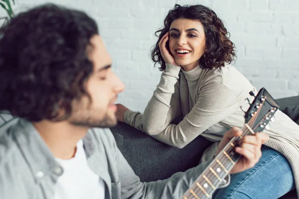 Novio tocando la guitarra y cantando para la novia sonriente en casa - foto de stock