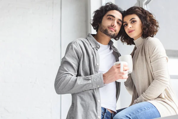 Joven pareja sosteniendo tazas de café y mirando a la cámara en casa - foto de stock