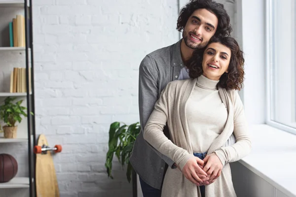 Lächelndes junges Paar, das sich umarmt und zu Hause in die Kamera schaut — Stockfoto