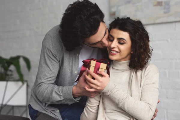 Boyfriend presenting gift to girlfriend and kissing her at home — Stock Photo