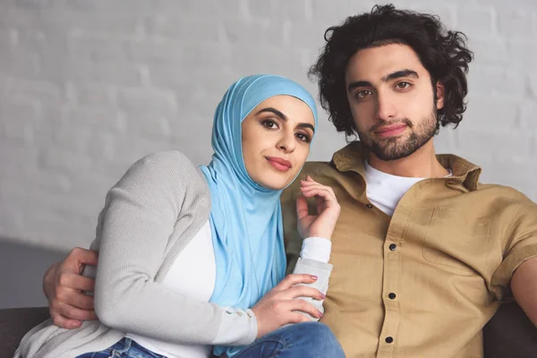 Feliz pareja musulmana sentado en el sofá en la sala de estar y mirando a la cámara - foto de stock
