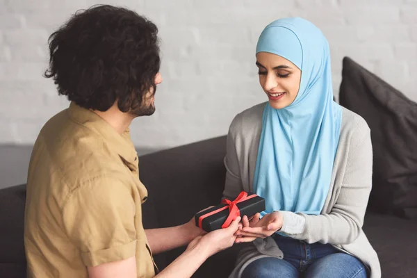 Muslim boyfriend presenting gift box to girlfriend in hijab in living room — Stock Photo