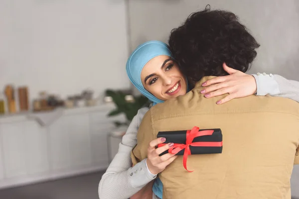 Muslim girlfriend in hijab hugging boyfriend and holding present at home — Stock Photo