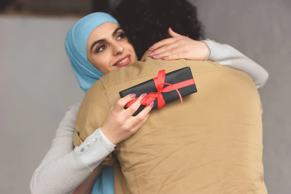 Muslim girlfriend in hijab holding present box and hugging boyfriend at home — Stock Photo