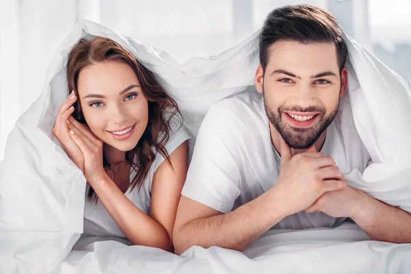 Portrait of young happy couple covered with blanket looking at camera — Stock Photo