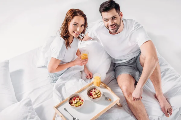 Высокий угол зрения молодой пары с завтраком на деревянный поднос глядя на камеру в постели — стоковое фото