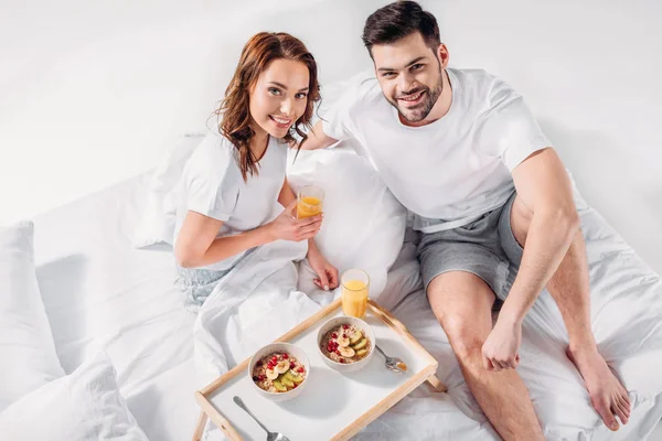 High angle view of young couple with breakfast on wooden tray looking at camera in bed — Stock Photo