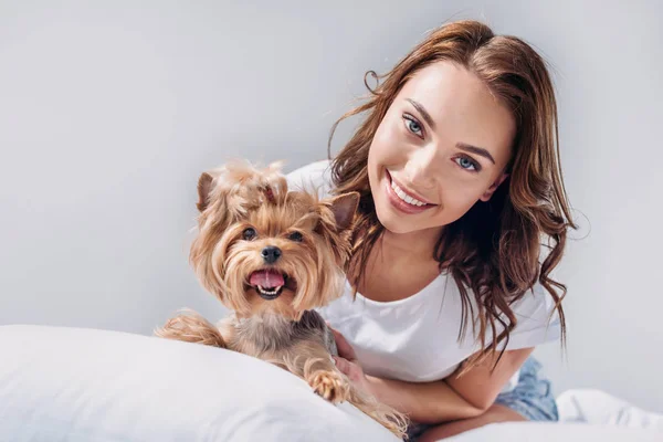 Portrait of young smiling woman with yorkshire terrier resting on bed isolated on grey — Stock Photo