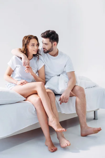 Jeune homme étreignant petite amie tout en étant assis sur le lit ensemble isolé sur gris — Photo de stock