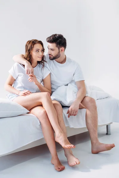 Young man hugging girlfriend while sitting on bed together isolated on grey — Stock Photo