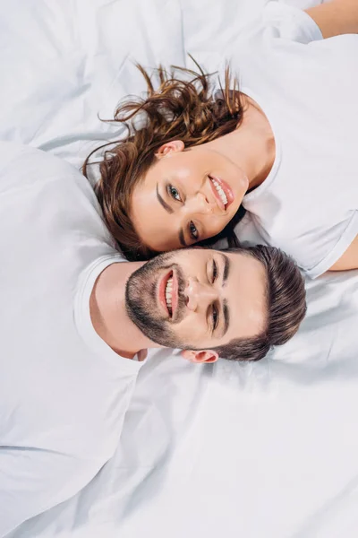 Вид сверху на молодую улыбающуюся влюбленную пару, которая смотрит в камеру, лежа вместе на кровати — стоковое фото