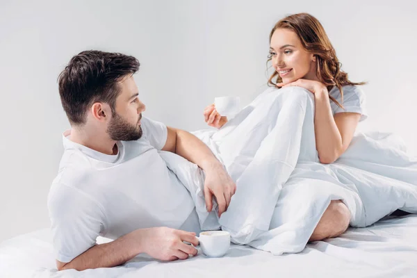 Jeune couple avec des tasses de café reposant au lit ensemble isolé sur gris — Photo de stock
