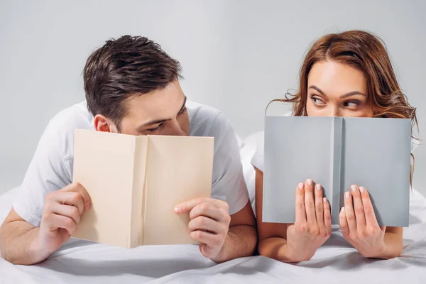 Obscured view of young couple covering faces with books while lying on bed isolated on grey — Stock Photo