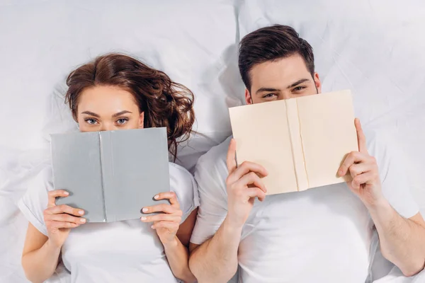 Vista aérea de la joven pareja cubriendo caras con libros mientras está acostada en la cama - foto de stock