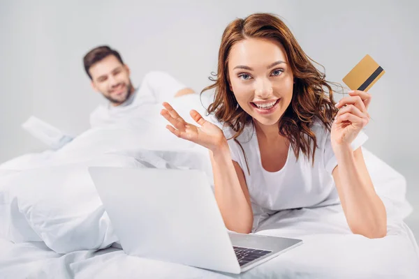Enfoque selectivo de la mujer sonriente con tarjeta de crédito y portátil acostado en la cama junto con el novio aislado en gris - foto de stock