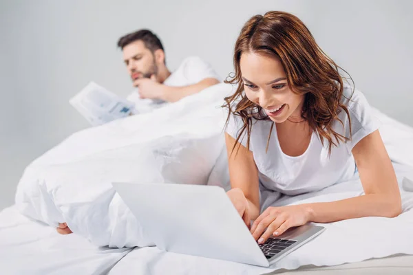 Foco seletivo de mulher sorridente usando laptop enquanto deitado na cama junto com o namorado isolado no cinza — Fotografia de Stock