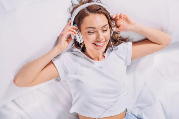 Overhead view of young smiling woman listening music in headphones in bed — Stock Photo