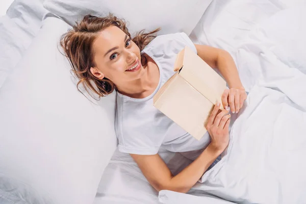 Вид зверху усміхненої жінки з книгою, що дивиться на камеру, лежачи в ліжку — стокове фото