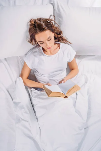 Overhead view of focused young woman reading book in bed — Stock Photo