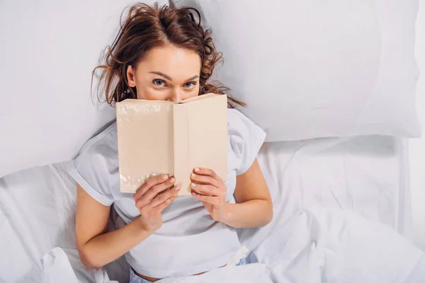 Vista oscura de la mujer cubriendo la cara con libro y mirando a la cámara mientras está acostado en la cama - foto de stock