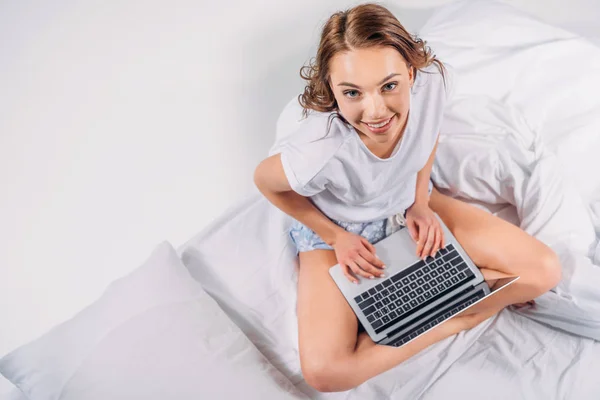 Над головой вид улыбающейся женщины с ноутбуком, сидящим на кровати — стоковое фото