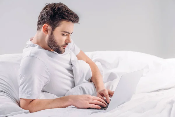 Vista laterale di focalizzato uomo barbuto utilizzando computer portatile a letto isolato su grigio — Foto stock