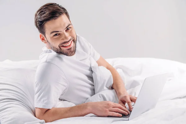 Портрет усміхненого чоловіка з ноутбуком відпочиває в ліжку ізольовано на сірому — стокове фото