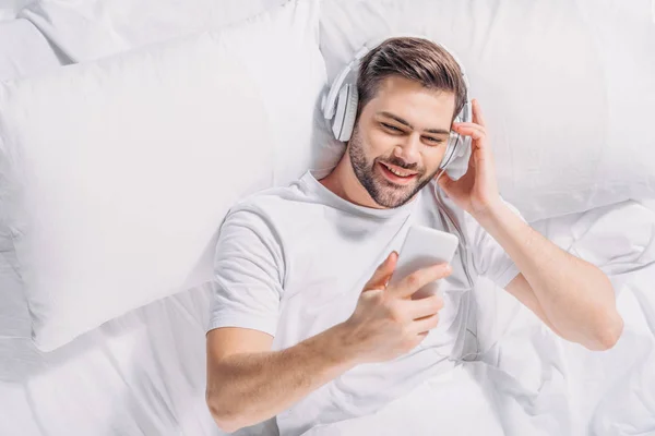 Vista aérea del hombre sonriente en auriculares usando teléfono inteligente en la cama - foto de stock