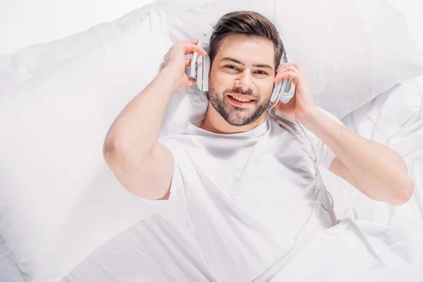 Vista aérea del hombre sonriente escuchando música en auriculares mientras descansa en la cama - foto de stock