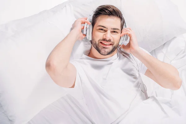 Vue aérienne d'un homme souriant écoutant de la musique dans un casque pendant qu'il se repose au lit — Photo de stock