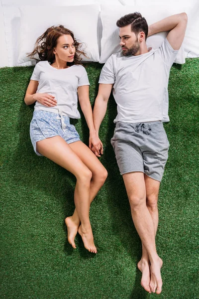 Vista aérea de pareja joven cogida de la mano mientras está acostada sobre almohadas sobre hierba verde - foto de stock
