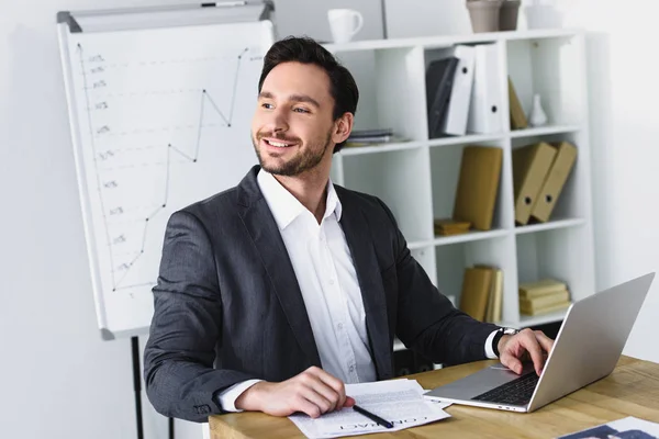 Lächelnder gutaussehender Geschäftsmann, der am Tisch sitzt und im Büro wegschaut — Stockfoto