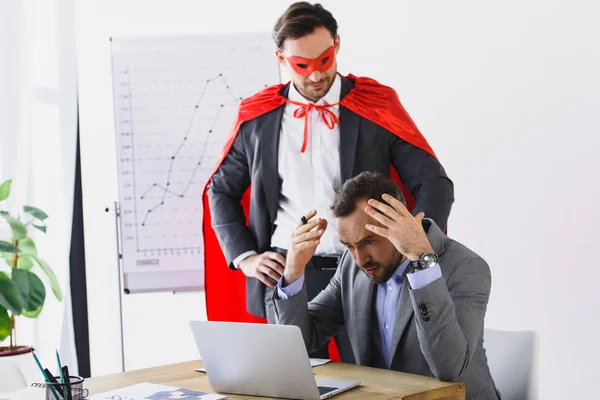 Super hombre de negocios en máscara y capa ayudando a hombre de negocios enojado con el trabajo en la oficina - foto de stock