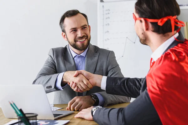 Super businessman in mask and cape shaking hands with smiling businessman in office — Stock Photo