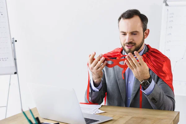 Super homme d'affaires en cape regardant masque rouge au bureau — Photo de stock