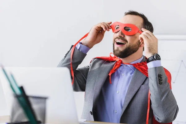 Excitado súper hombre de negocios en capa mirando a través de máscara roja en la oficina - foto de stock