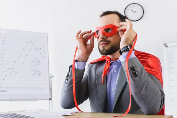 Super hombre de negocios en capa mirando a través de máscara roja en la oficina - foto de stock