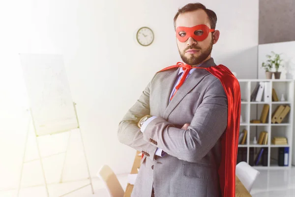 Serio super hombre de negocios en máscara y capa de pie con los brazos cruzados en la oficina - foto de stock