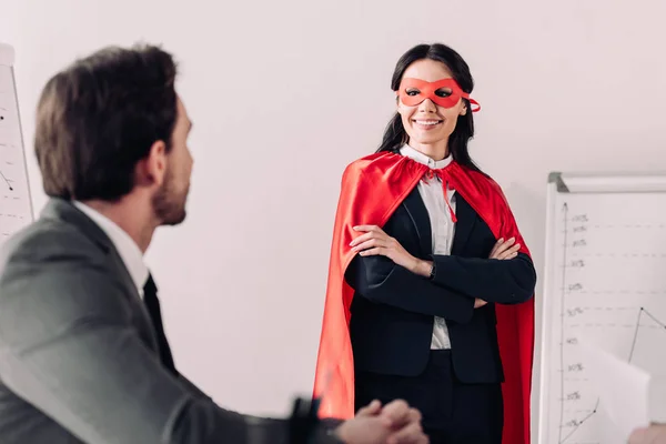 Sonriente super mujer de negocios en máscara y capa mirando a hombre de negocios en la oficina - foto de stock