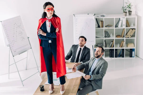 Super mujer de negocios en máscara y capa de pie sobre la mesa en la oficina - foto de stock