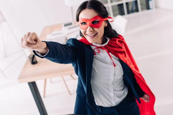 Attractive super businesswoman in cape and mask standing with hand up in office — Stock Photo