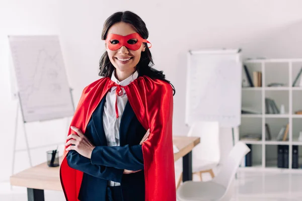 Attrayant super femme d'affaires en cape et masque debout avec les bras croisés dans le bureau — Photo de stock