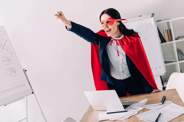 Attractive super businesswoman in cape and mask screaming and standing with hand up in office — Stock Photo