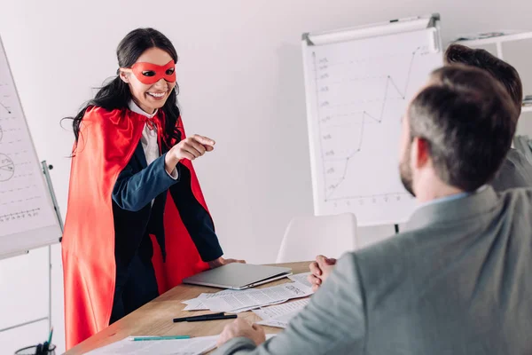 Sonriente super mujer de negocios en máscara y capa apuntando a los hombres de negocios en la oficina - foto de stock