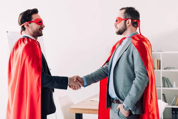Side view of handsome super businessmen in masks and capes shaking hands in office — Stock Photo